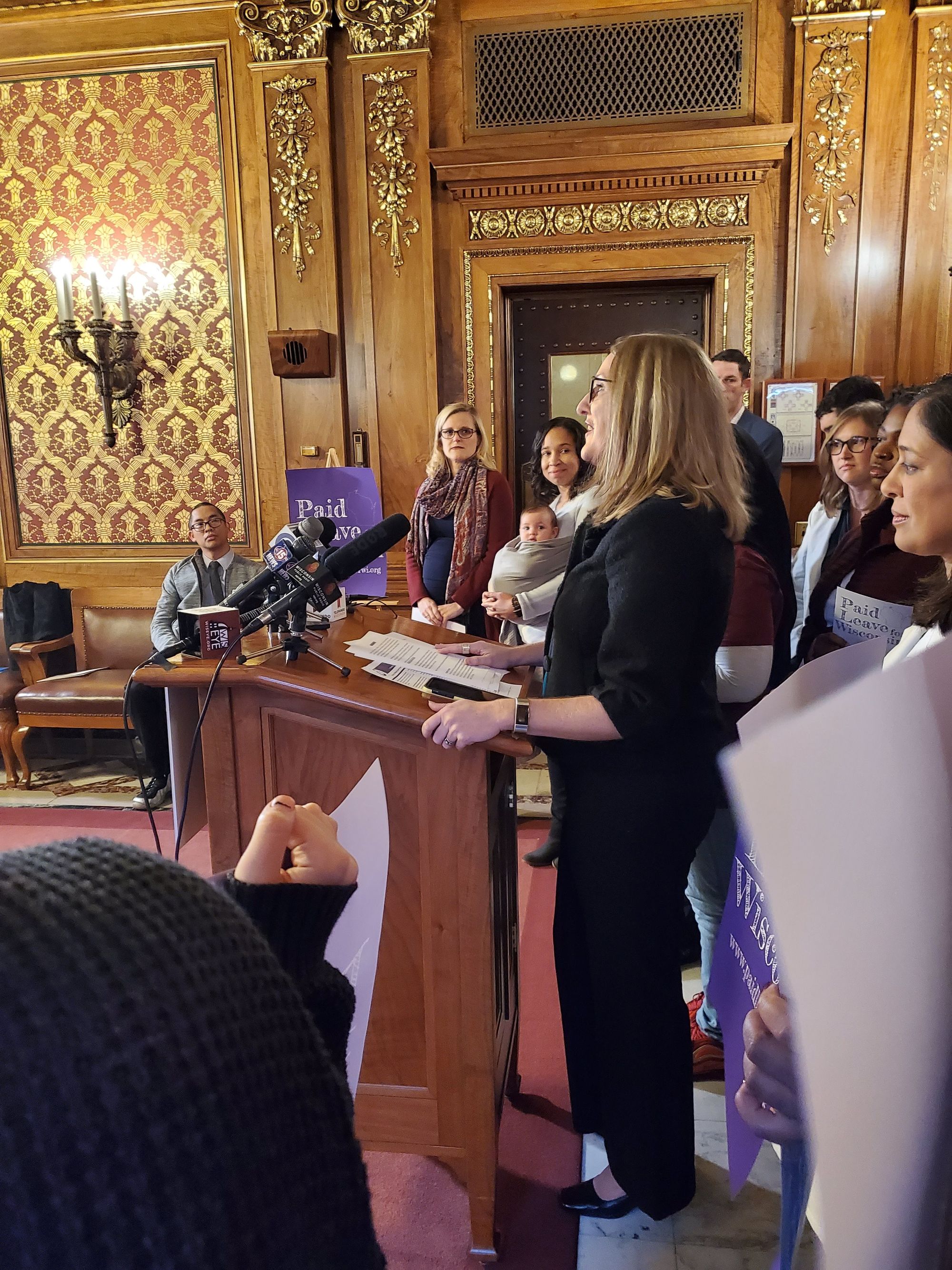 Image of a light skinned woman with light brown hair and dark glasses speaking at a wood podium. There are a variety of people standing around her, some with purple signs that say Paid Leave Wisconsin. .